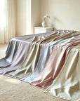 A bed draped with multiple TENCEL™ Lyocell Bed Sheet in soft neutral tones including gray, champagne, blush pink, and cream, styled in a bright, minimalist bedroom with a small white side table and vase.