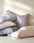 A styled collection of pastel-colored pillows, including light blue, dusty purple, and soft pink, arranged on a bed with warm sunlight casting shadows on the wall.