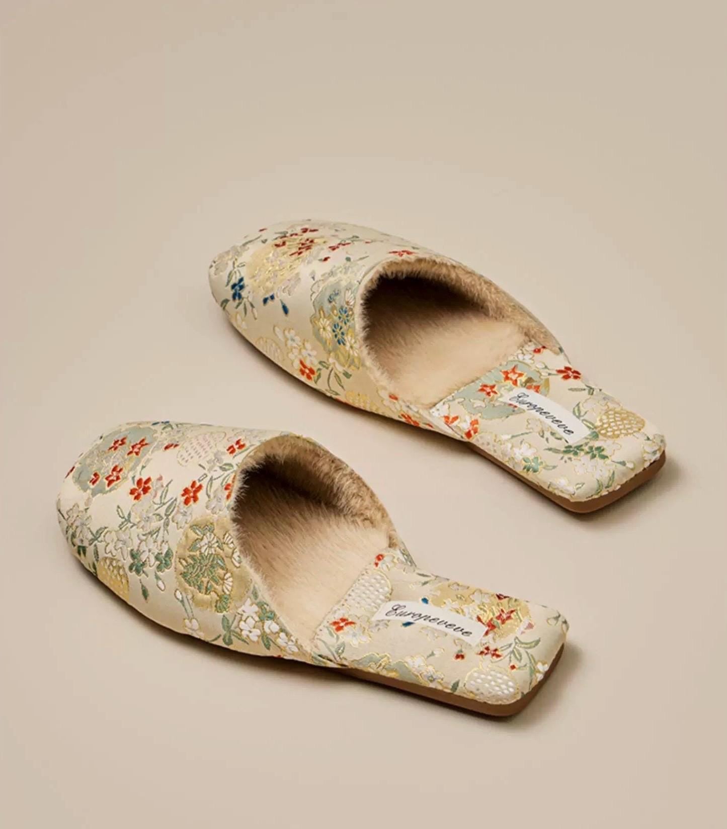 Pair of beige floral slippers with fluffy cream-colored lining. The slippers have a square toe and feature a delicate floral pattern in shades of orange, green, and white. The brand name 'Kungeneene' is subtly printed on the insole. These comfortable-looking house slippers are perfect for relaxing at home.