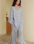 May include: A light blue pajama set. The top has a ruffled collar and button-up front. The long sleeves have gathered cuffs. The pants are wide-legged. The fabric appears soft and lightweight. The outfit is displayed on a person standing in a room with a neutral-colored wall