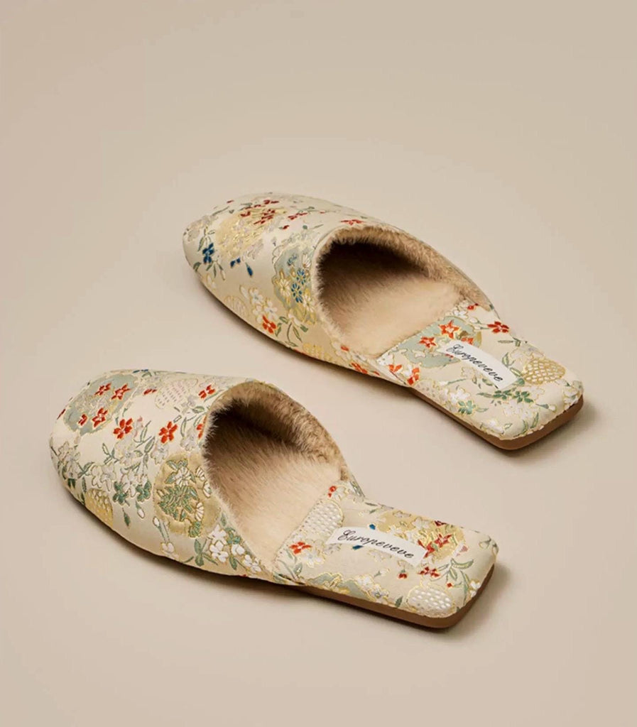 Pair of beige floral slippers with fluffy cream-colored lining. The slippers have a square toe and feature a delicate floral pattern in shades of orange, green, and white. The brand name &#39;Kungeneene&#39; is subtly printed on the insole. These comfortable-looking house slippers are perfect for relaxing at home.