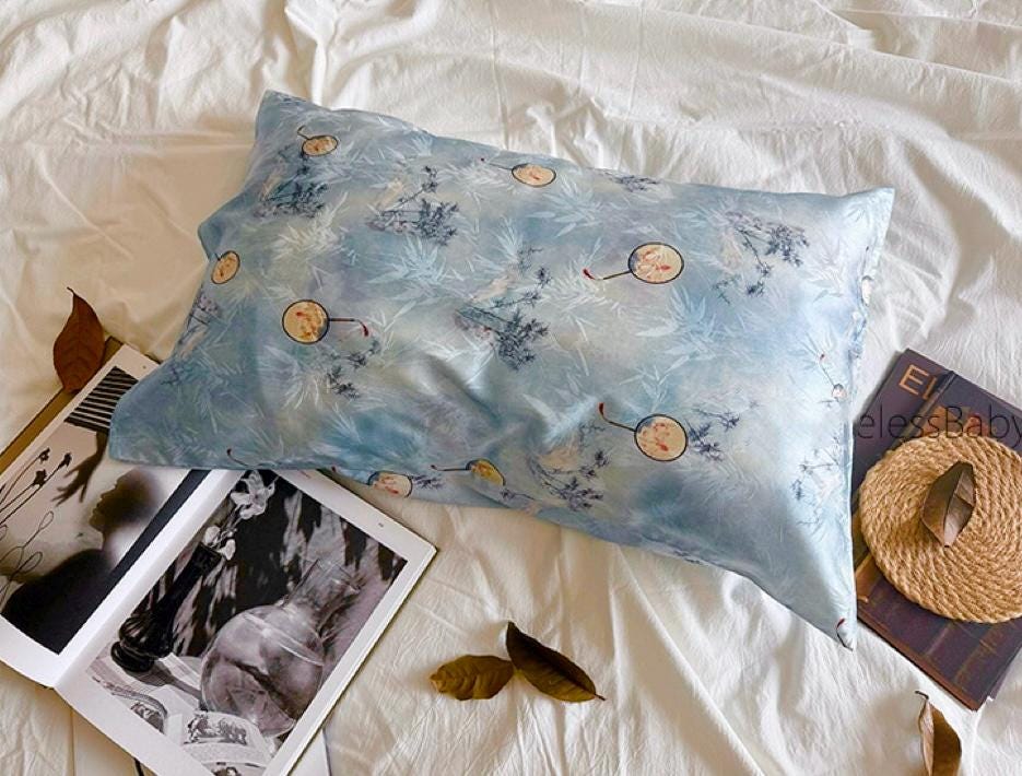 A soft sky blue silk pillowcase on a bed, featuring an artistic design of round Chinese hand-fan motifs and ink-painted landscape patterns.