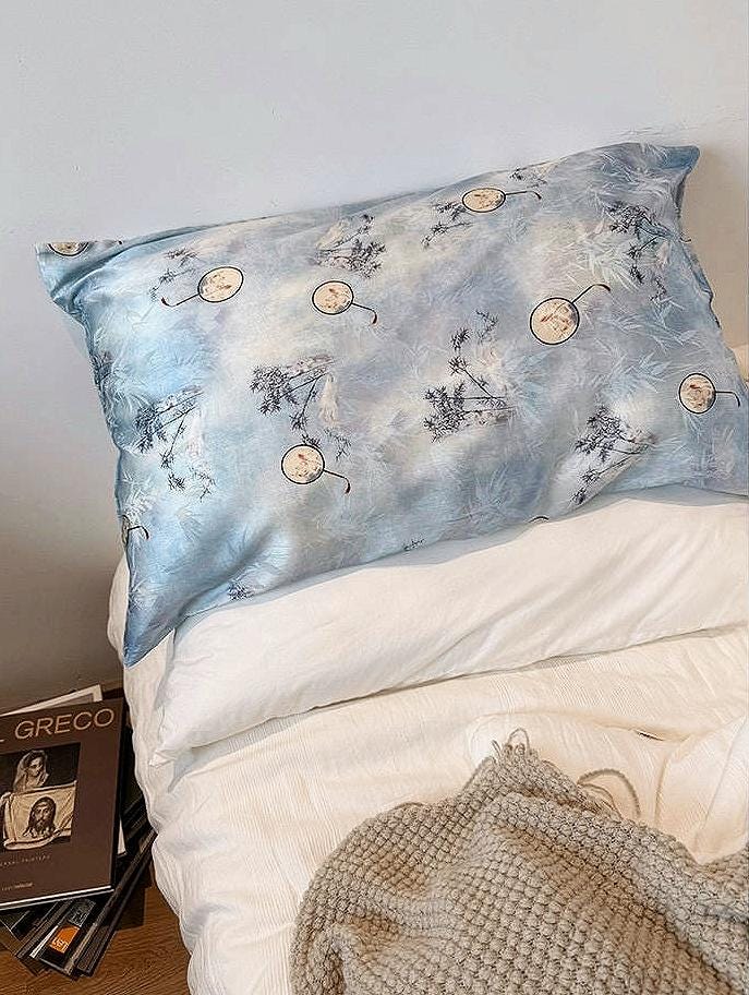 A soft sky blue silk pillowcase on a bed, featuring an artistic design of round Chinese hand-fan motifs and ink-painted landscape patterns.