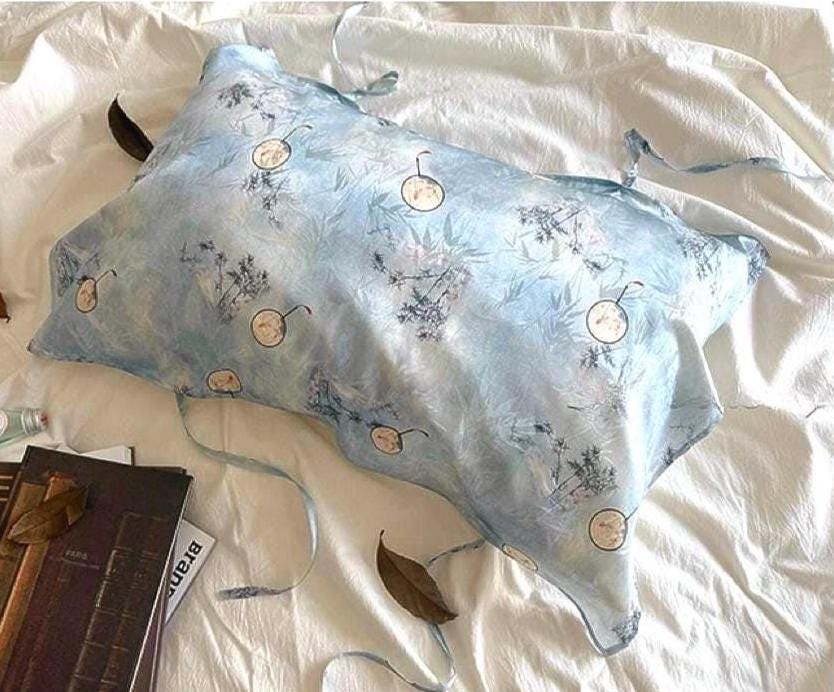 A soft sky blue silk pillowcase on a bed, featuring an artistic design of round Chinese hand-fan motifs and ink-painted landscape patterns.