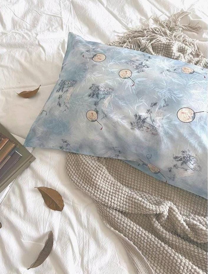 A soft sky blue silk pillowcase on a bed, featuring an artistic design of round Chinese hand-fan motifs and ink-painted landscape patterns.