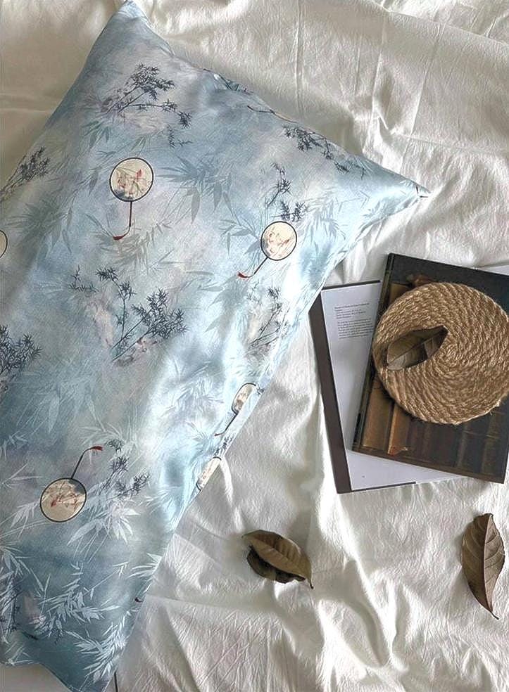 A soft sky blue silk pillowcase on a bed, featuring an artistic design of round Chinese hand-fan motifs and ink-painted landscape patterns.