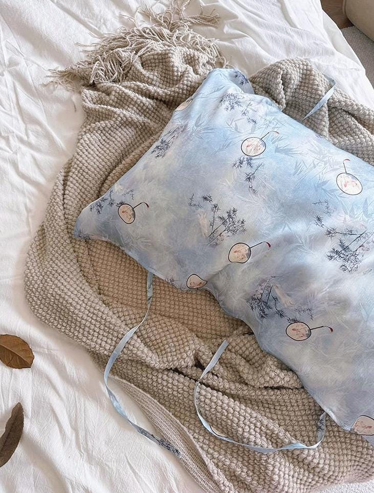 A soft sky blue silk pillowcase on a bed, featuring an artistic design of round Chinese hand-fan motifs and ink-painted landscape patterns. silk pillow cases