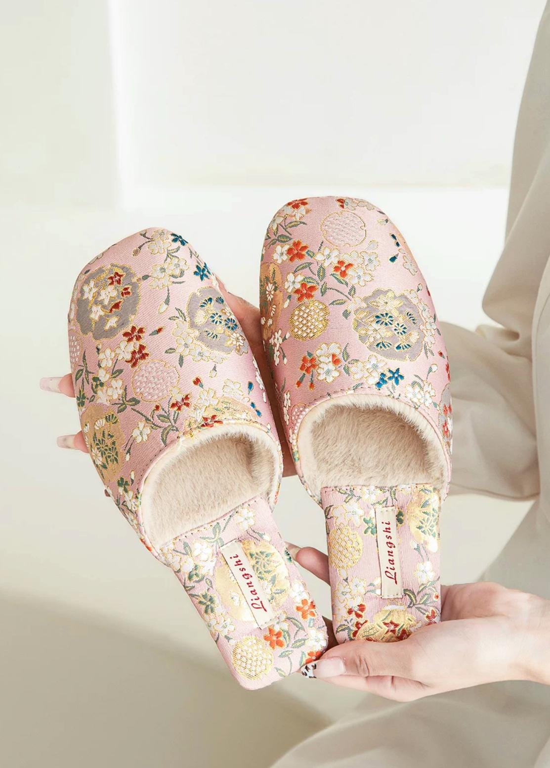 Pink floral slippers with a plush, light beige interior. The slippers feature an embroidered floral pattern in shades of pink, orange, and gray. The brand name 'Liangshi' is embroidered on the insole. These comfortable and stylish slippers are perfect for relaxing at home.