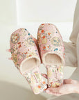 Pink floral slippers with a plush, light beige interior. The slippers feature an embroidered floral pattern in shades of pink, orange, and gray. The brand name &